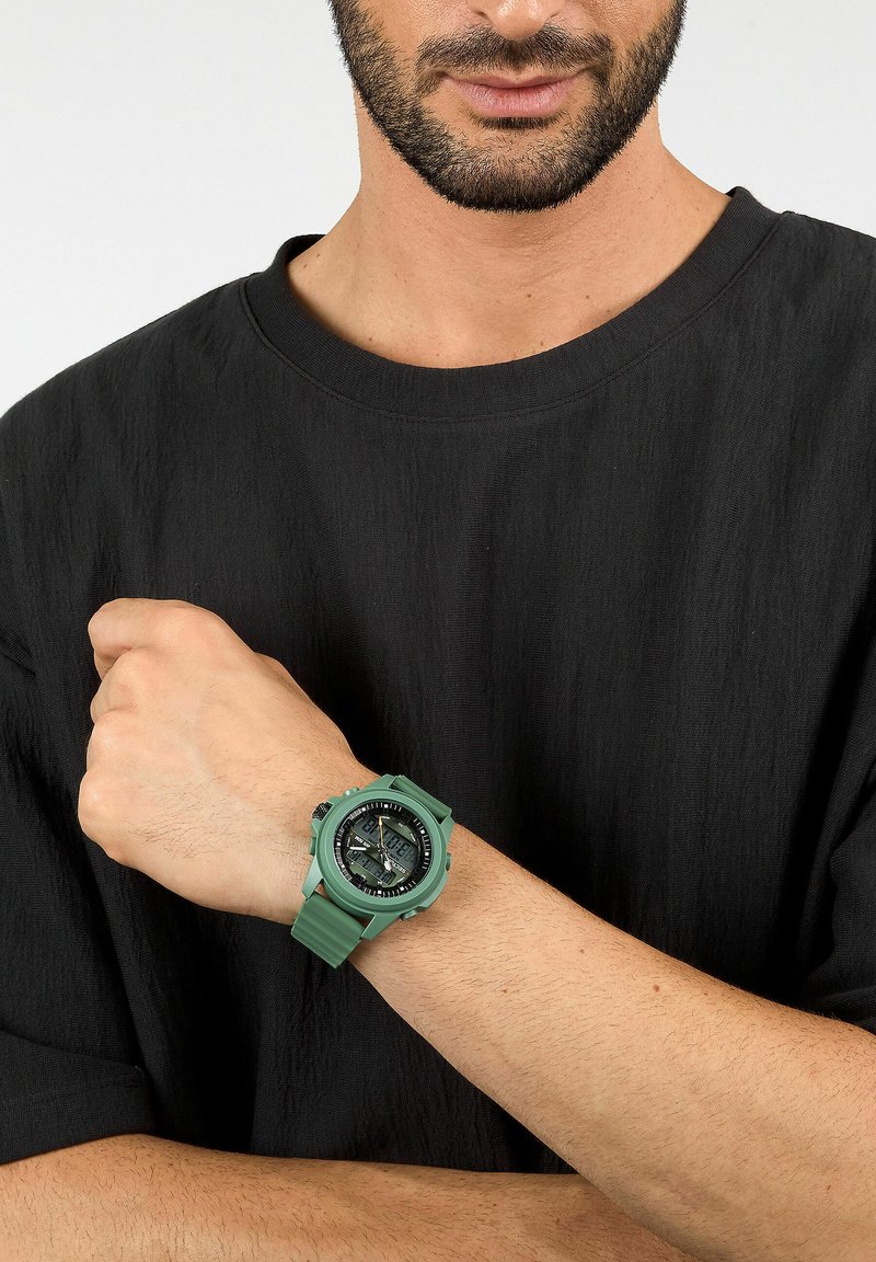 Orologio in gomma verde con quadrante rotondo, display analogico e digitale, cinturino texturizzato e quadrante nero a contrasto, indossato su una mano contro una camicia nera.