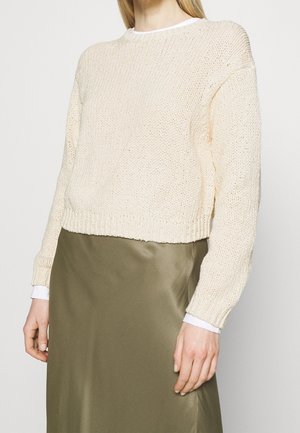 Vrouw draagt een crèmekleurige, gebreide cropped trui over een wit overhemd en een olijfgroene satijnen rok, staand met ontspannen armen.