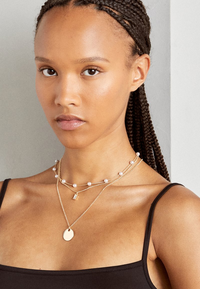 Kerangka kalung emas bertumpuk dengan aksen mutiara, pendant geometris, dan pesona bundar, dipakai di atas atasan tanpa lengan berwarna hitam. Tekstur kulit yang halus.