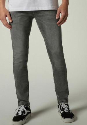 Mężczyzna w szarych jeansach typu slim fit, czarno-białych trampkach i białej koszulce, stojący z rękami swobodnie opuszczonymi wzdłuż ciała.