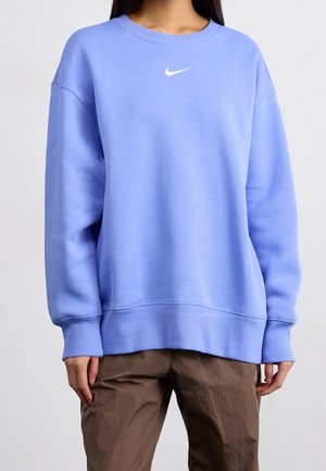 Osoba ubrana w jasnoniebieską bluzę Nike i brązowe spodnie, stojąca na tle jednolitego jasnego tła.