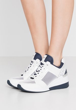 Hvide og marineblå kilehøje sneakers med meshpaneler, snørebånd og mærket stof på hælen, båret på bare fødder mod en lys baggrund.