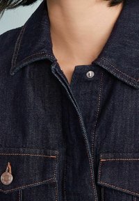 Koszula dżinsowa w ciemnoniebieskim kolorze z ostro zakończonym kołnierzykiem, przednim zapięciem na napy oraz dwiema kieszeniami na piersi. Charakteryzuje się kontrastowymi pomarańczowymi przeszyciami.