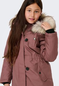 Rózsaszín, bélelt parkaszövet, műszőrme kapucnival, gombos záródással, derékban megkötővel és oldalsó zsebekkel. Sima textúrájú anyag.