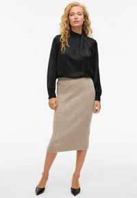 Blusa de satén negro con cuello alto y mangas largas, combinada con una falda lápiz de punto beige, complementada con tacones negros de punta.
