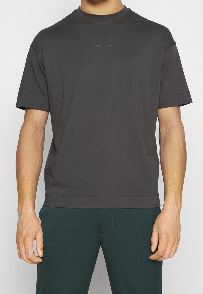 Šedé bavlněné tričko s kulatým výstřihem, krátkými rukávy a decentním textovým logem "Emporio Armani" na přední straně. Hladká textura, uvolněný střih.