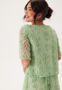 Top trasparente di colore verde chiaro con applicazioni floreali 3D, maniche corte e taglio dritto. Texture liscia con motivi floreali sparsi.