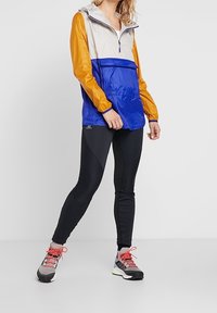 Färgglad vindjacka med en design i beige, blått och orange; i kombination med svarta leggings och flerfärgade löparskor.