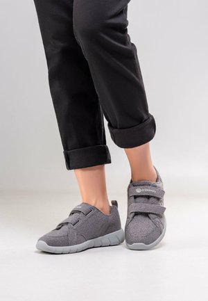 Stopy w czarnych podwiniętych spodniach i szarego materiałowe trampki na rzepy na jasnoszarym tle.