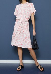 Różowa sukienka z krótkim rękawem w kwiatowy wzór, z przodem na guziki i paskiem do zawiązania; stylizowana z czarnymi sandałami na ramiączkach i małą czarną torebką.