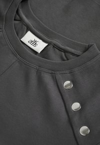 Tumehall sweatshirth, millel on ümar kaelus. Vasakul õlal on hõbedased metallnööbid ning kaeluses on kangaslabel.