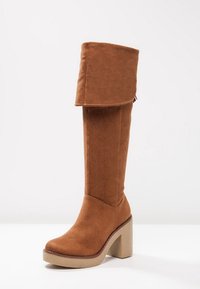 Botte haute jusqu'au genou en daim marron avec un revers plié, semelle plateforme beige épaisse et bout rond. Lignes épurées et texture lisse.