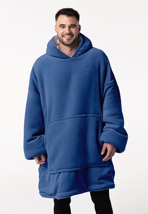 Lächelnder Mann, der einen übergroßen blauen Fleece-Hoodie mit großer Fronttasche und Kapuze trägt, steht vor einem schlichten weißen Hintergrund.