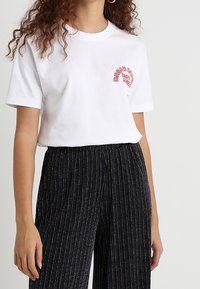 Camiseta de algodón blanca con mangas cortas, con texto rojo en un diseño curvado. Combinada con pantalones amplios de pierna ancha, negros, texturizados y plisados.