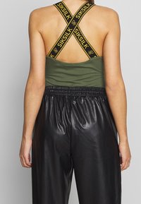Canotta verde con spalline incrociate nere con branding giallo, abbinata a pantaloni in pelle nera lucida a vita alta.