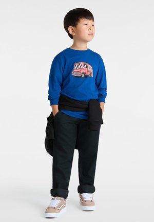 Blauwe lange mouwen shirt met een rode voedingstruck grafiek, zwarte broek en tan sneakers. Zwarte sweatshirt om de taille geknoopt. Witte achtergrond.