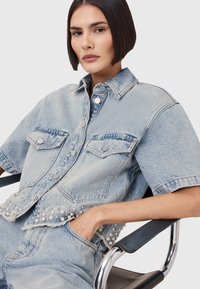 Lichtblauwe denimjas met korte mouwen, zilveren knoopdetails, twee voorkwabben en parelversieringen langs de zoom.