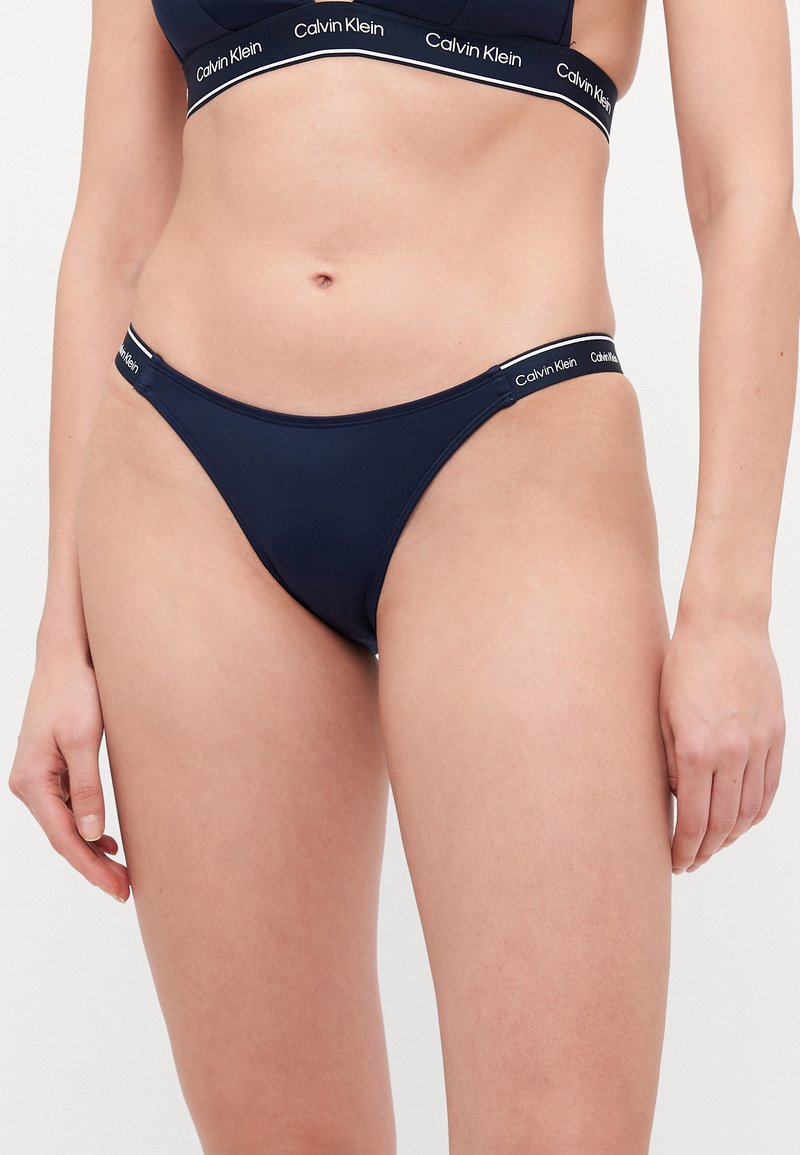 Sötétkék bikini alsó, minimális takarással. Simább szövetből készült, Calvin Klein logós derekú, kontrasztos fehér részletekkel.