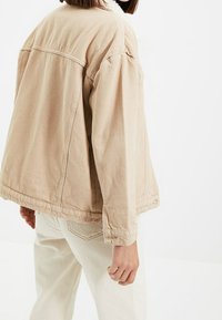 Beige džínsová bunda s volným střihem, viditelným šitím a měkkým, texturovaným límcem. Spárovaná se světlými kalhotami.