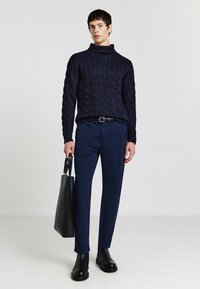 Maglione a collo alto intrecciato navy, abbinato a pantaloni navy e stivaletti neri alla caviglia. La modella tiene in mano una borsa tote nera elegante. Sfondo semplice.