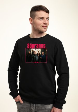Homme portant un sweat-shirt noir avec une image du casting de The Sopranos et un texte rouge, regardant vers sa droite avec une main dans la poche.