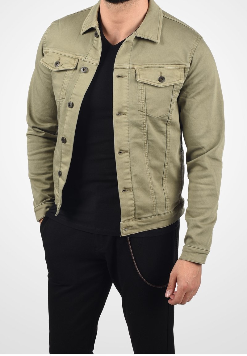 Homme portant une veste boutonnée vert olive sur une chemise noire avec un pantalon noir, une main dans la poche, sur un fond blanc.