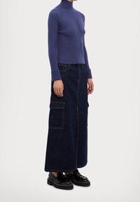Paarse geribde turtleneck met lange mouwen en een voorzak, gecombineerd met een donkere denim maxi-rok met zijzakken en gestikte naden. Zwarte platform schoenen.
