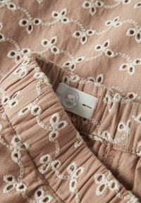 Brūns audums ar dekoratīvu baltu izšuvumu ar ziedu rakstiem. Ir savilkts jostasvietā un aizdare ar plastmasas pogu.