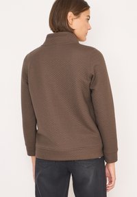 Pullover marrone, texturizzato, con colletto alto e maniche lunghe. Presenta un orlo aderente e polsini a coste. Indossato sopra jeans scuri.