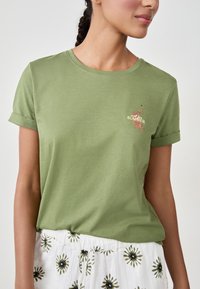 Frau trägt ein olivgrünes T-Shirt mit hochgekrempelten Ärmeln, das mit einem Leopardenmuster und dem Text "SUMMER" verziert ist, kombiniert mit weißen, floral gemusterten Jogginghosen mit Kordelzug.