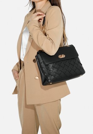Borsa nera con texture intrecciata, hardware dorato e tracolla a catena. Ha una forma strutturata, abbinata a un giubbotto tan e una camicia bianca.