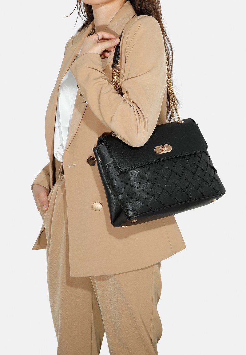 Borsa nera con texture intrecciata, hardware dorato e tracolla a catena. Ha una forma strutturata, abbinata a un giubbotto tan e una camicia bianca.