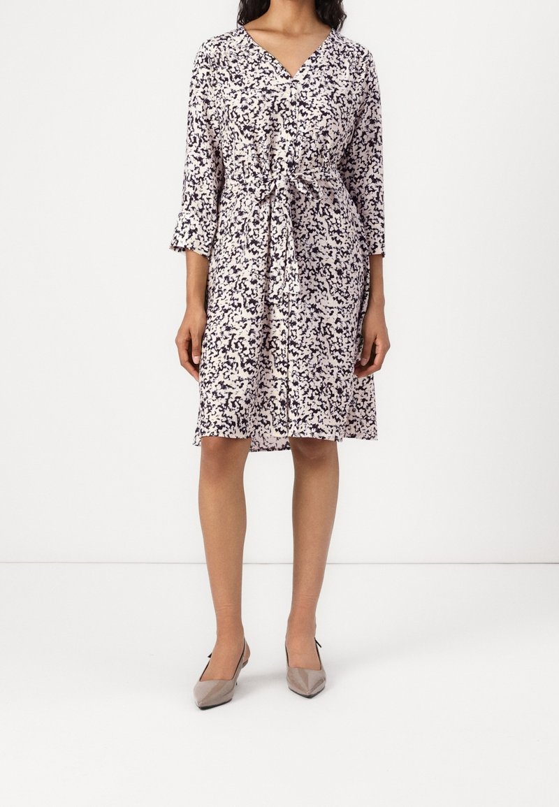 Kukallinen mekko, jossa on 3/4 -hihaiset kellohihat, V-pääkallo ja sidottava vyötärö. Pääasiassa kermaista väriä, jossa on tummansinisiä abstrakteja kuvioita. Yhdistetty beigen värisiin tasapohjaisiin kenkiin.