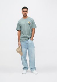 Jauns vīrietis stāv, valkājot pelēku grafisko T-kreklu, gaiši zilas, brīvas piegriezuma džinsas, baltas sporta kurpes, tur kreisajā rokā bēša cepuri, neitrāla fona priekšā.