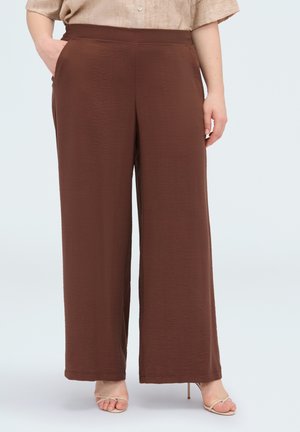 Femme portant un pantalon marron taille haute à jambes larges, associé à des sandales à talons hauts beiges à brides, sur un fond uni.