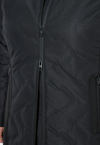 Svart texturerad jacka med dubbla dragkedjor fram och sidoficka, närbild på torso och armar.