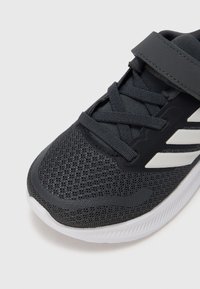 Zapato deportivo negro con parte superior de malla, rayas blancas contrastantes y cordones. Cuenta con un cuello acolchado y una suela texturizada para mejor agarre.