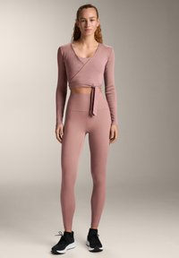 Roza dolgotrajna majica z dolgimi rokavi in detajlom za vezanje ter gamaše z visokim pasom, obe iz mehkih, raztegljivih materialov. Črne športne superge dopolnjujejo obleko.