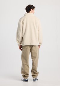 Beige fleece jacket with a high collar, relaxed fit, and textured exterior, paired with light beige trousers and gray sneakers.