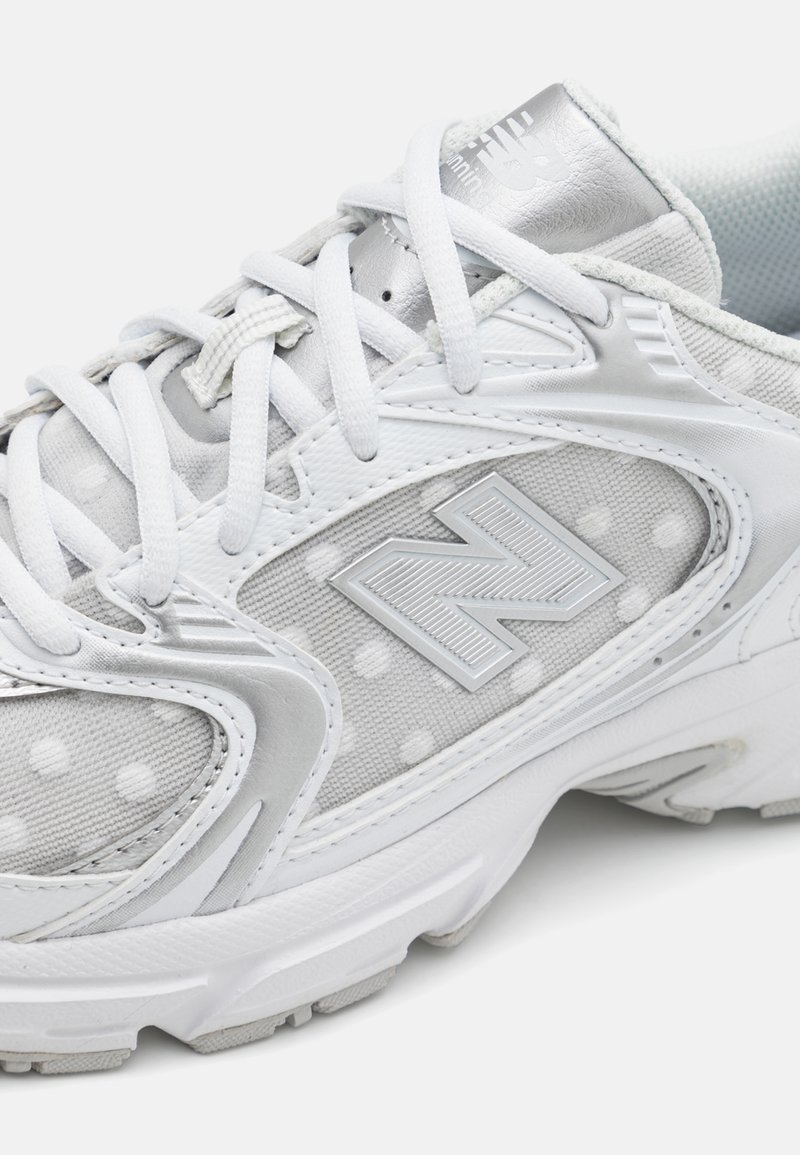 Baskets de sport blanches avec une texture à pois, des accents argentés et un logo 'N' bien en vue. Lacets ronds et talon rembourré pour le confort.