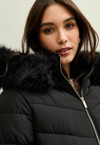 Młoda kobieta w czarnej kurtce puchowej z kapturem wykończonym futrem, częściowo zapięta, łagodnie patrzy w obiektyw na neutralnym tle.