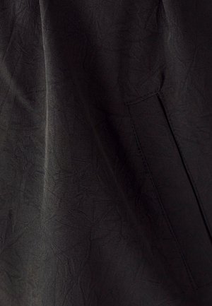 Črn teksturiran material z subtilnimi vzorci in detajlom stranske žepne. Material se zdi lahek z gubanimi zaključki.