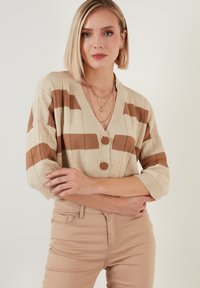 Beige pruhovaný kardigan s hnědými pruhy, hlubokým V-výstřihem, s dvěma knoflíkovými zapínáními a tříčtvrtečními rukávy, v kombinaci se světle béžovými přiléhavými kalhotami.