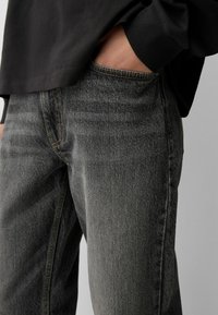 Persona che indossa jeans in denim nero sbiadito e una maglietta nera a maniche lunghe con una mano nella tasca anteriore.