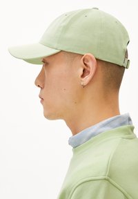 Casquette en coton vert avec un rebord incurvé, couronne structurée et trous de ventilation. Portée avec une chemise à col bleu clair et un sweat-shirt vert.