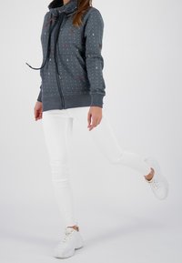 Felpa grigia con zip e motivo a pois, colletto alto e dettagli in pelle, abbinata a jeans skinny bianchi e sneakers bianche.