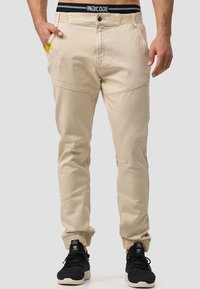 Pantalones beige con un diseño ajustado, puños elásticos, bolsillos delanteros y detalles de costura, combinados con zapatillas negras.