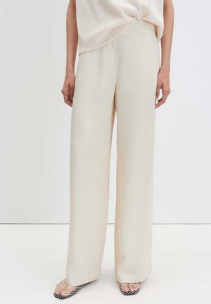 Femme vêtue d’un pantalon crème à jambes larges et d’un haut sans manches beige clair, debout devant un fond blanc uni.