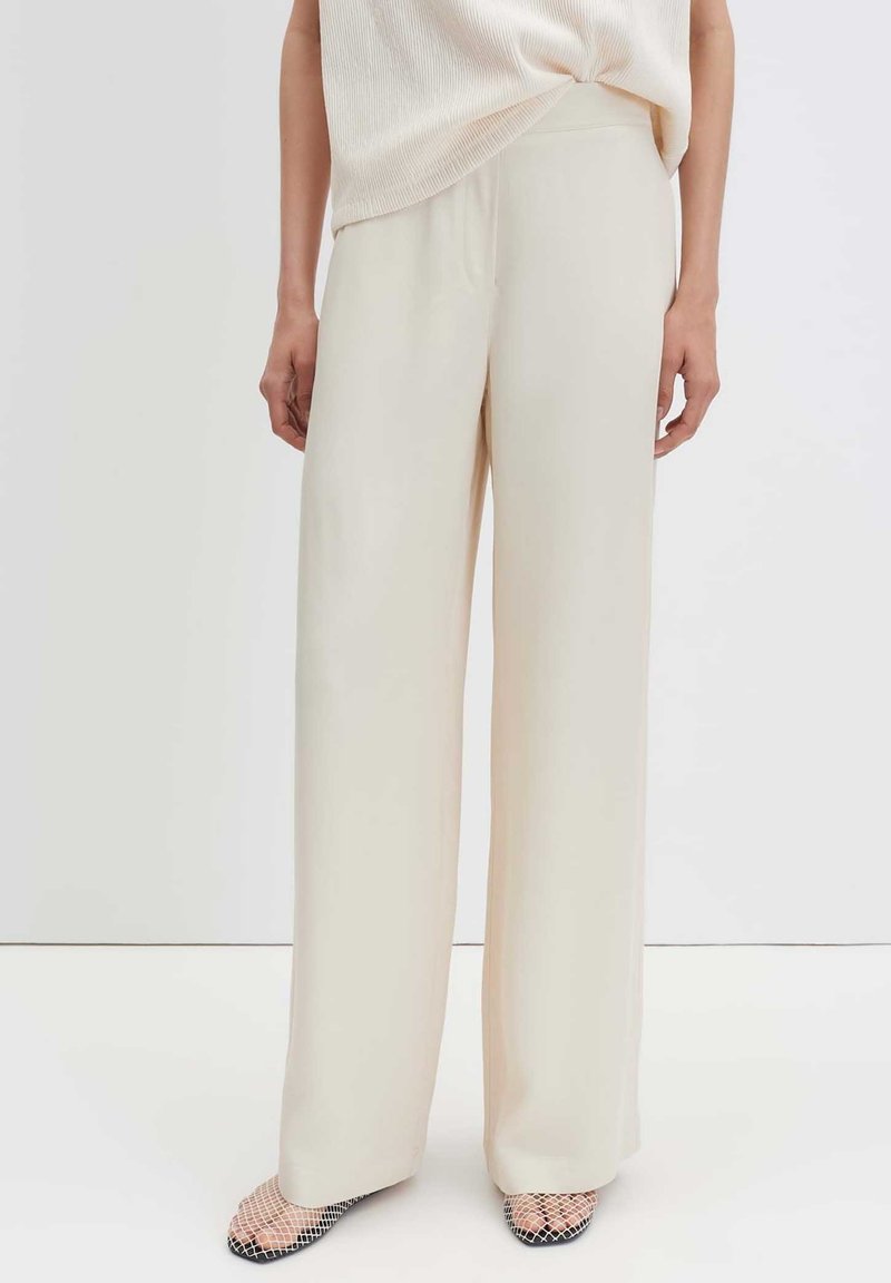 Woman wearing cream wide-leg trousers and a light beige sleeveless top, standing against a plain white background.