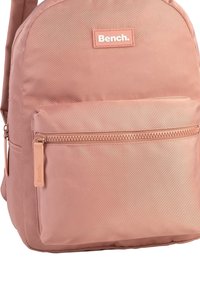 Sac à dos en nylon rose de forme arrondie, avec une poche zippée à l'avant et un patch logo rectangulaire. Texture lisse et fermeture éclair robuste.